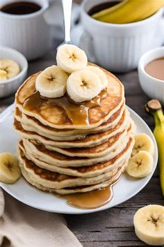 stack of pancakes with syrup and bananas on a white plate next to two cups of coffee