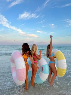 three women in bikinis are standing on the beach with large rafts and one woman is holding her hand up