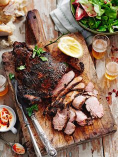 a wooden cutting board topped with meat next to a bowl of salad and two glasses of beer