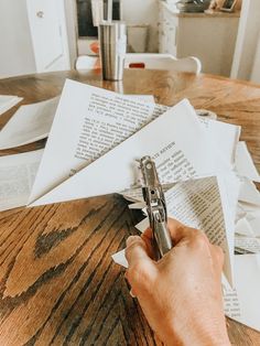 a person holding a pen over some papers on a wooden table in front of an open book