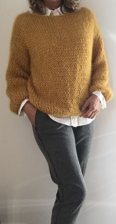 a woman standing in front of a white wall wearing a yellow sweater and grey pants