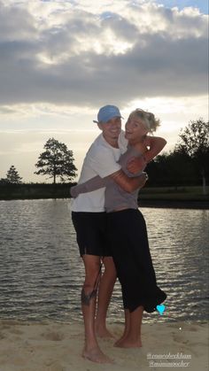 a man and woman hugging on the beach at sunset with clouds in the sky behind them