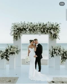 a bride and groom standing in front of an arch with flowers on it at the beach