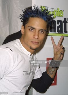 a man with blue hair and piercings making the peace sign in front of a white wall