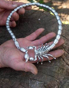 a person holding a silver scorpion necklace on top of a piece of wood with another hand