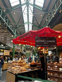 people are shopping at an outdoor market with red umbrellas over the food stalls and tables