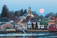 a full moon rises over a small town on the water's edge with houses in the foreground