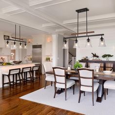 a dining room table and chairs with lights hanging from the ceiling above it in front of an open kitchen area