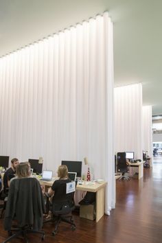 two people sitting at desks in an office with white drapes on the wall