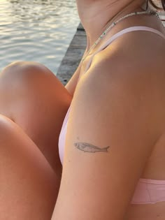 a woman with a small fish tattoo on her arm sitting next to the water and looking off into the distance