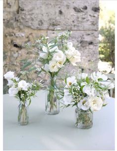 three clear vases with white flowers in them on a blue table cloth near an old brick wall