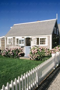 a white picket fence in front of a house with pink flowers on the bushes next to it