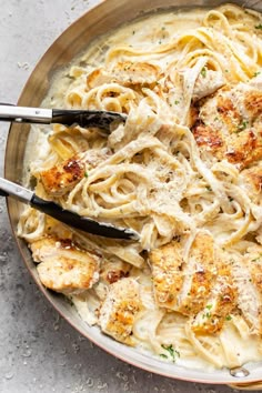 a bowl filled with pasta and chicken on top of a white countertop next to two tongs