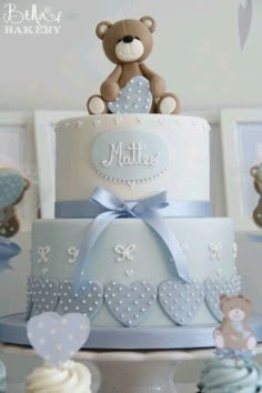 a teddy bear sitting on top of a blue and white tiered cake next to cupcakes