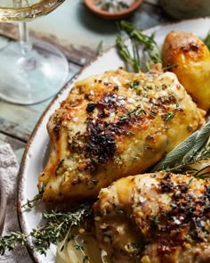 two pieces of chicken on a plate with potatoes and herbs next to a glass of wine