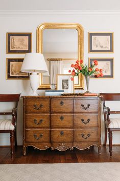 a dresser with flowers on it in front of a mirror and framed pictures above it