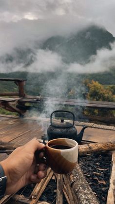 a person holding a coffee cup in front of a campfire with mountains in the background