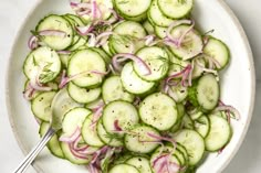 a white bowl filled with cucumbers and onions next to a silver serving spoon