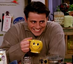 a man holding a yellow smiley face mug in front of some jars and cups on a table