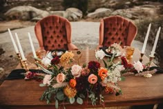 a wooden table topped with lots of flowers next to two red velvet chairs covered in candles