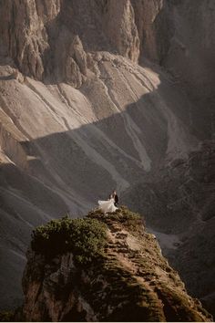 a bride and groom are standing on the edge of a cliff overlooking a valley with mountains in the background