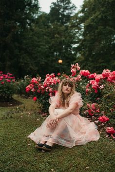 a woman in a pink dress sitting on the ground next to some flowers and bushes