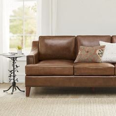 a brown leather couch with two pillows on it in front of a white wall and window