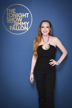 a woman with long red hair standing in front of a blue wall wearing a black jumpsuit