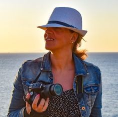 a woman holding a camera and smiling at the ocean with her hat over her head