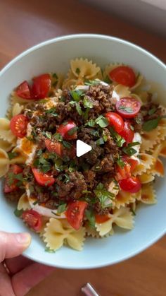 a person is holding a bowl of pasta with meat and tomatoes on the table next to a fork