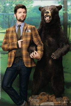 a man standing next to a giant bear in front of a forest backdrop with trees