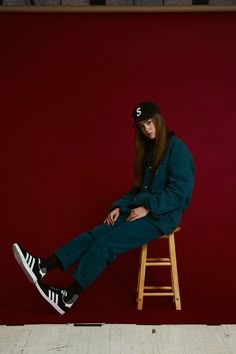 a woman sitting on top of a wooden stool wearing a black hat and green pants