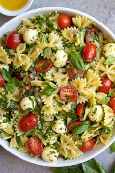 a white bowl filled with pasta salad next to a cup of olive oil and a spoon