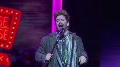 a man singing into a microphone in front of a neon sign that reads peace please