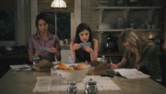 three women sitting at a kitchen table looking at their cell phones and eating oranges