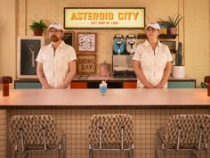 two men standing behind a counter at a fast food restaurant in front of a neon sign that reads asteroid city