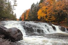 a waterfall in the woods surrounded by rocks and trees with fall foliage on it's sides