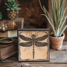 a wooden block with a dragonfly on it next to a potted plant and books