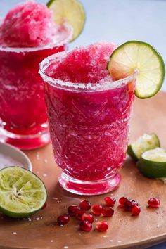 two glasses filled with red and green drink next to limes on a cutting board