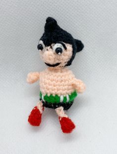 a small crocheted doll is posed on a white surface with red and green feet