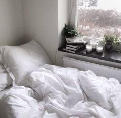 an unmade bed with white sheets and pillows next to a window sill filled with books