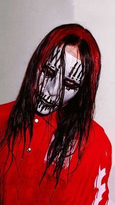 a man with long hair and painted face wearing a red shirt is standing in front of a white wall