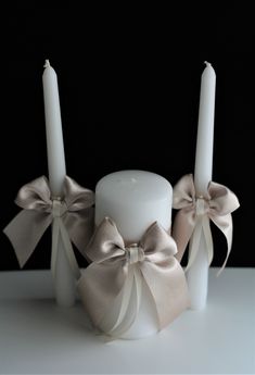 two white candles with bows on them sitting in front of a black background and one candle is lit