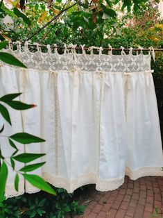 a white curtain hanging from a clothes line in front of some bushes and trees with green leaves