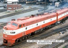 a red and white train traveling down train tracks next to a loading platform with buildings in the background