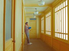 a man standing in an empty hallway holding a book and looking out the window at something