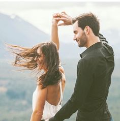 a man standing next to a woman on top of a mountain with her hair blowing in the wind