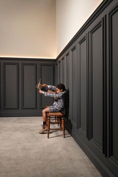 a man sitting on top of a wooden chair next to a tall black paneled wall