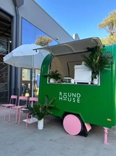 a green food truck parked in front of a building with pink chairs and an umbrella