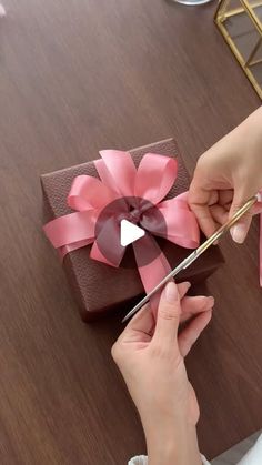 a woman is cutting a pink ribbon on a brown box with a pair of scissors